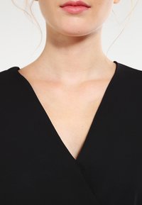 Svart omlottklänning med V-ringning, slät textur och korta ärmar, som lyfter fram ringningen och nyckelbenet.