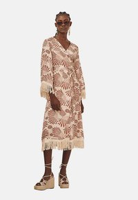 Vestido maxi color crema con patrones florales rojos, mangas con flecos y dobladillo. Combinado con sandalias de plataforma color beige y gafas de sol oversized.