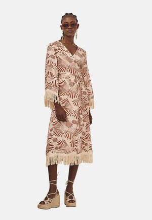 Vestido maxi color crema con patrones florales rojos, mangas con flecos y dobladillo. Combinado con sandalias de plataforma color beige y gafas de sol oversized.