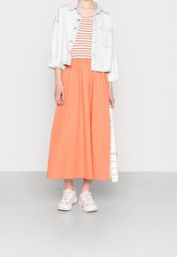 Veste en denim sur un haut rayé rouge et blanc, associée à une jupe midi orange et des baskets blanches. L'utilisateur tient une ceinture blanche avec des clous.