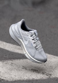 Grauer Laufschuh mit einem strukturierten Mesh-Obermaterial, weißer Sohle und subtilen blauen Akzenten. Verfügt über ein Schnürdesign und eine geschwungene Ferse.