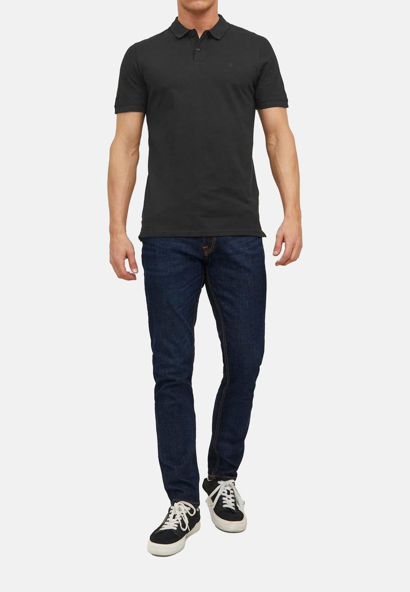 Schwarzes Poloshirt mit Kragen, kombiniert mit dunkelblauen Jeans und schwarzen Sneakers. Das Shirt hat ein kleines Logo auf der Brust.