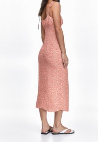 Mujer con un vestido midi sin mangas de flores naranjas y tirantes atados, usando sandalias planas blancas, de pie frente a un fondo blanco.