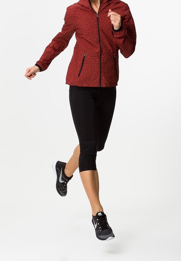 Personne portant une veste rouge, des leggings courts noirs et des chaussures de course Nike noires en plein milieu d’une foulée sur un fond blanc uni.