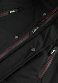 Veste en tissu noir avec des accents de fermeture éclair rouge, des boutons-pression argentés et le logo "Clarks" sur deux tirettes de fermeture éclair. Texture lisse et design ergonomique.