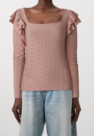 Długi rękaw w kolorze różanym z kwadratowym dekoltem, falbanką na ramionach i teksturowaną tkaniną z wzorami ażurowymi. Połączony z jasnoniebieskimi dżinsami.