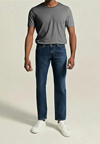 Homme portant un t-shirt gris à manches courtes, un jean bleu foncé et des chaussures blanches sans lacets, debout sur un fond clair uni.