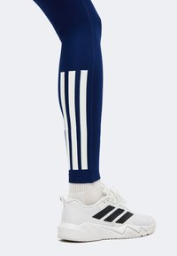 Jambe portant un legging bleu foncé avec trois bandes blanches et une chaussure de sport blanche avec trois bandes noires sur un fond clair.
