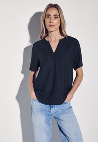 Schwarzes, kurzärmliges Oberteil mit V-Ausschnitt, glatter Textur und lockerer Passform, kombiniert mit hellblauen Jeans. Weicher Stoff, minimalistisches Design.