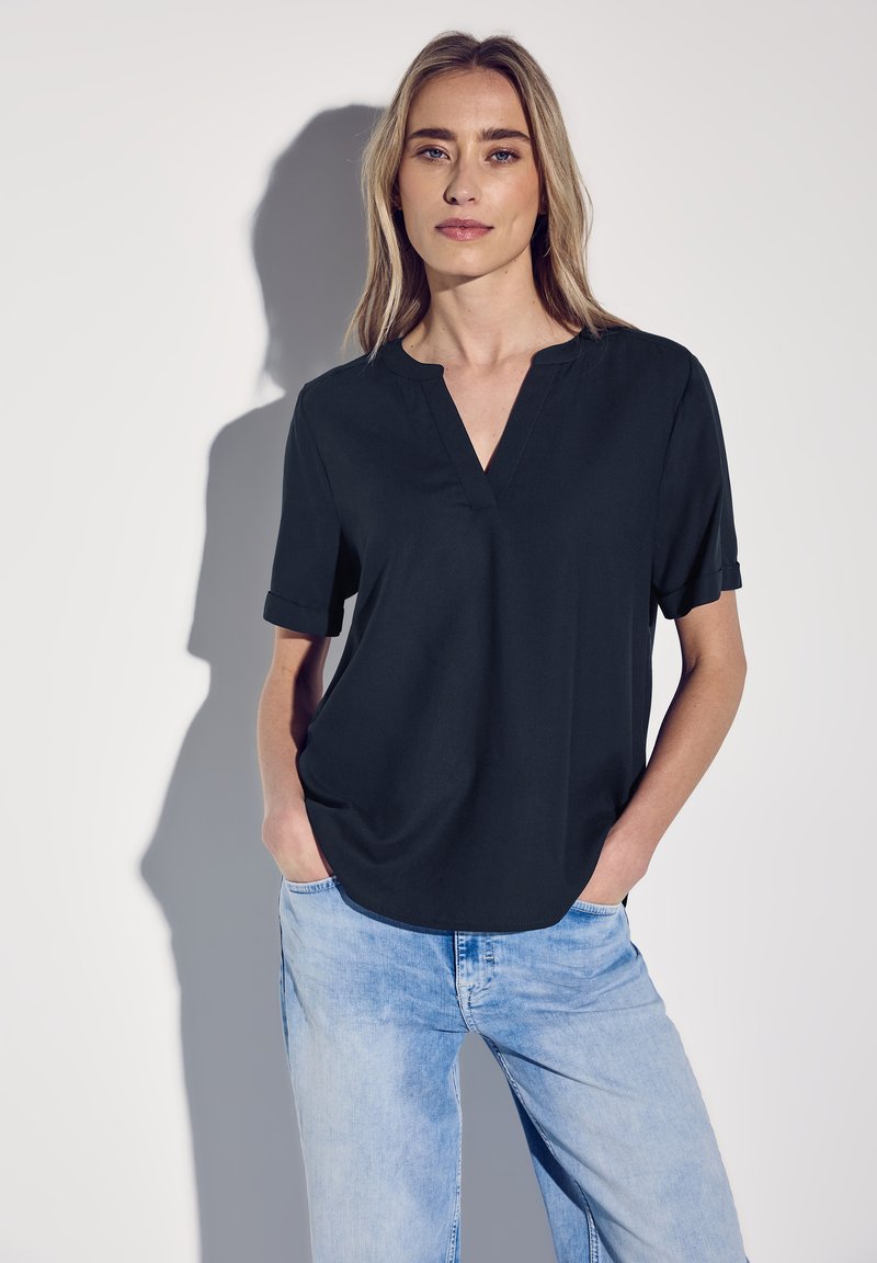 Schwarzes, kurzärmliges Oberteil mit V-Ausschnitt, glatter Textur und lockerer Passform, kombiniert mit hellblauen Jeans. Weicher Stoff, minimalistisches Design.