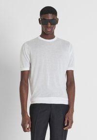 Suéter de punto blanco de manga corta con cuello redondo y puños acanalados. Se lleva con pantalones negros ajustados y gafas de sol rectangulares negras.