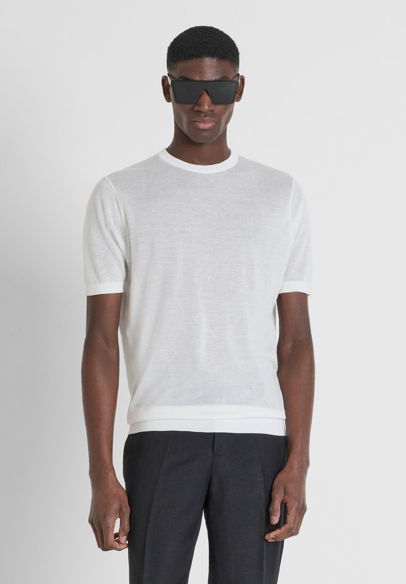 Suéter de punto blanco de manga corta con cuello redondo y puños acanalados. Se lleva con pantalones negros ajustados y gafas de sol rectangulares negras.