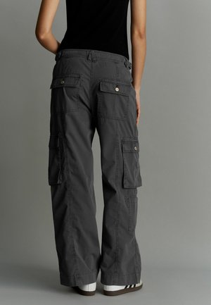 Graue Cargo-Hosen mit mehreren Taschen und goldfarbener Hardware, in lockerer Passform und mit weichem Stoffgefühl, kombiniert mit weißen Sneakers.
