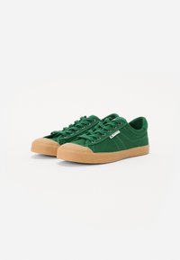 Green canvas sneakers with a gum rubber sole, rounded toe cap, and a low-profile design. Features lace-up closure and a side logo tag.