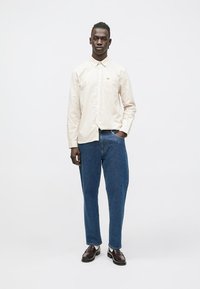 Beige-ruudullinen pitkähiha-paita, jossa on kaulus, yhdistettynä löysiin sinisiin farkkuihin ja tummanruskeisiin kenkiin, seisoo valkoista taustaa vasten.