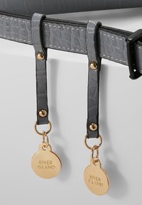 Ceinture grise texturée avec deux sangles pendantes, chacune ornée d'un charme circulaire doré gravé "RIVER ISLAND."