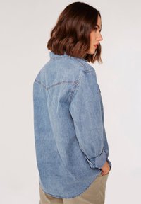 Lys blå denimskjorte med knap-ned krave, oprullede ærmer og synlige stikninger, der har et afslappet fit og buet kant.