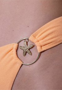 Perzikkleurige bikini band met een zachte textuur, voorzien van een ronde gouden metalen ring en een accent in de vorm van een zee ster.