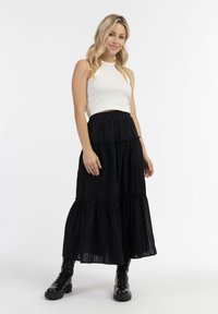 Zwarte gelaagde maxi rok met een gestructureerde stof, gecombineerd met een witte geribde tanktop. Het model draagt zwarte veterschoenen.