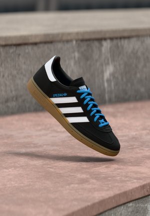 Svart Adidas Spezial-sneakers med hvite striper, blå lisser og gummisåle, plassert over en betongflate mot en bakgrunn av steinvegg.