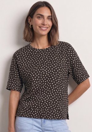 Smilende kvinde med skulderlangt brunt hår iført gyldne øreringe, en sort kortærmet top med hvide polkaprikker og lyseblå jeans.