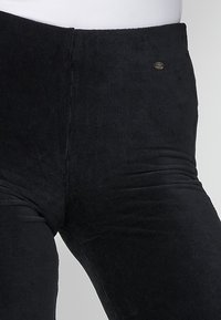 Pantalon en velours côtelé noir taille haute avec un petit logo métallique rond sur la hanche droite, porté sur un torse de mannequin blanc.