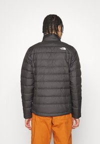 Giacca a vento nera con design trapuntato, colletto alto e logo sulla schiena. Indossata con pantaloni arancioni, che mostrano una trama traspirante.
