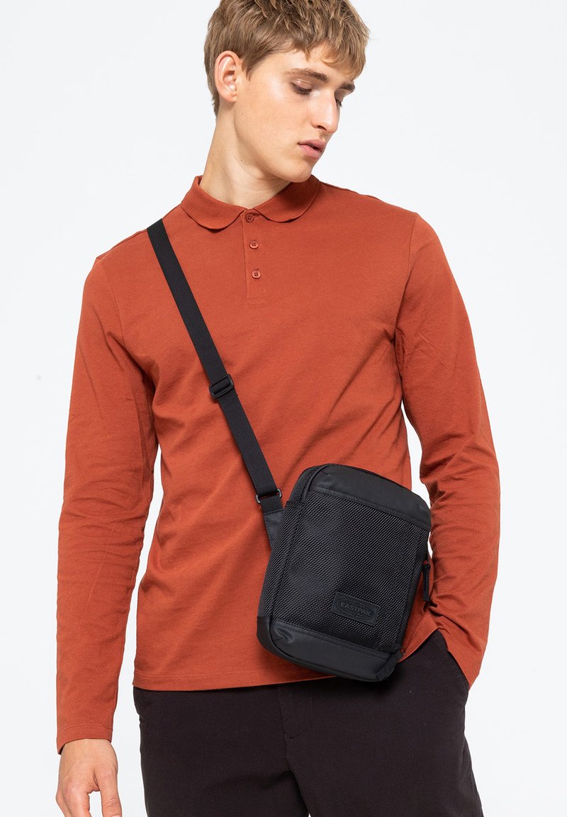 Borsa a tracolla nera con pannello anteriore in rete, forma rettangolare, tracolla regolabile e patch con logo. Indossata sopra una maglietta a maniche lunghe color ruggine.