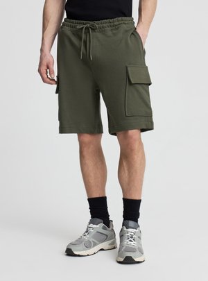 Mężczyzna w oliwkowych krótkich spodniach typu cargo z przednimi kieszeniami, czarnych skarpetkach i szarych butach sportowych stoi na tle jednolitego tła.