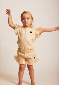 Helles gelbes Outfit mit Rüschärmeln und Shorts, versehen mit schwarzen Herzakzenten auf dem Shirt und den Shorts. Glattes, leichtes Material.