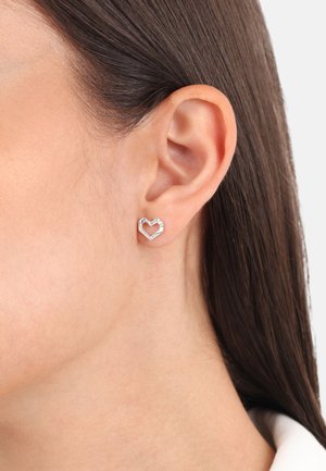 Pendientes de aro en forma de corazón en tono plateado con detalles geométricos y un acabado pulido, colocados en una oreja perforada.
