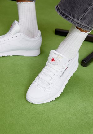 Valge sünteetiline toss, millel on perforatsioonidega varvas, Reebok'i kaubamärk ja iseloomulik logo. Kandnud ribakujulisi valgeid sokke rohelisel pinnal.