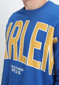 Sudadera azul de cuello redondo con gran texto amarillo que dice "HARLEM" y texto blanco más pequeño debajo. Material de algodón suave.