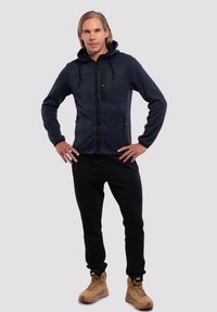 Marinblå zip-up hoodie med en svart panel och två sidfickor, parat med svarta joggers och bruna stövlar. Slät tygtextur.