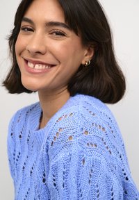 Pulover tricotat de culoare albastru deschis, cu o croială lejeră, având un model texturat cu ochiuri deschise și un gât larg, asortat cu cercei cercului aurii.