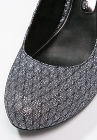 Zapato de tacón cerrado en color gris, con una tela de purpurina texturizada y un patrón de diamantes, forma redondeada y un interior negro suave.