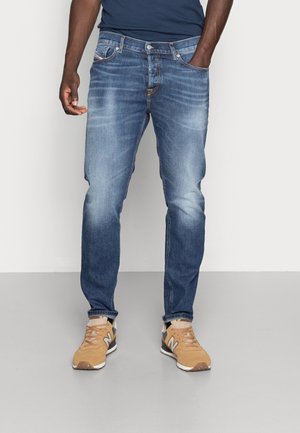 Mann, der blaue ausgewaschene Jeans und beige New Balance Sneakers trägt, steht vor einem einfarbigen hellgrauen Hintergrund.