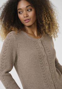 Beige knitted cardigan with a textured pattern and pearl buttons, featuring openwork detail along the sleeves. Round neckline.