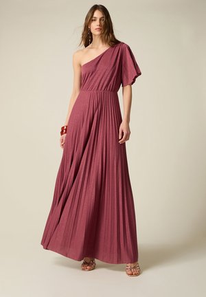 Frau trägt ein einseitig schulterfreies, plissiertes, bodenlanges Kleid in Mauve mit ausgestelltem Ärmel und Riemchensandalen vor einfachem Hintergrund.