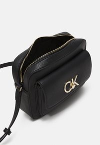 Borsa a tracolla in pelle nera texturizzata con zip dorata e logo CK in oro di Calvin Klein sulla patta della tasca anteriore.