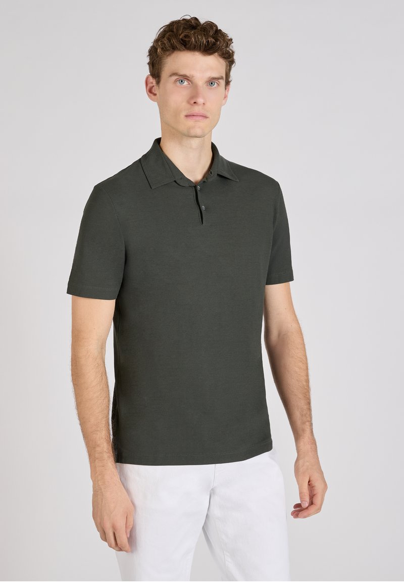 Polo de manga corta verde oscuro con cuello plano, tapeta de tres botones y textura suave, combinado con pantalones blancos.