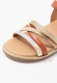 Sandały na paski z jasnobrązowej skóry, ze złotą brokatową podeszwą, irydyzującymi cekinami i dekoracyjną metalową płytką z logo.