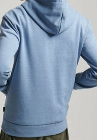 Persona que lleva una sudadera azul claro, vista desde atrás, con las manos en las caderas contra un fondo claro.
