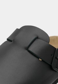 Svart lærklogg med matt overflate, med en bred stropp og en metallspenne. Innvendig foret med beigefarget semsket materiale.