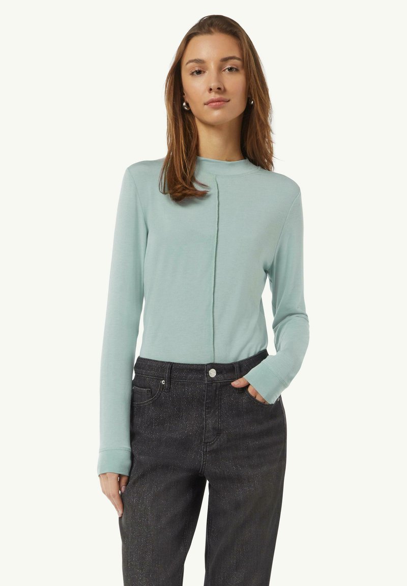 Hellblaues Langarmoberteil mit hohem Kragen und vertikalen Nahtdetails, kombiniert mit dunkel strukturierten, hoch taillierten Jeans.