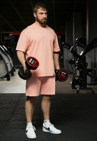 Completo sportivo in cotone rosa, composto da una maglietta a maniche corte e pantaloncini. La persona tiene in mano dei manubri neri con dettagli rossi, indossando sneakers bianche.