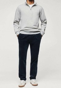 Sweat-shirt gris clair à fermeture éclair, pantalon bleu marine et baskets blanches avec semelles marron. La tenue présente un design décontracté et des textures douces.