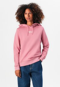 Hauts rose avec une poche avant et un capuchon à cordon. Présente un petit logo blanc sur la poitrine. Porté avec un jean en denim bleu.