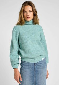 Maglione a collo alto a coste di un verde chiaro, con maniche lunghe. Texture morbida, vestibilità rilassata, abbinato a una gonna in denim blu.