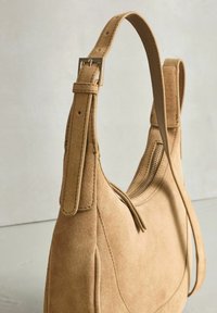 Bézs színű, sima tapintású velúr kézitáska, egy állítható pánttal és oldalt cipzáros nyílással.